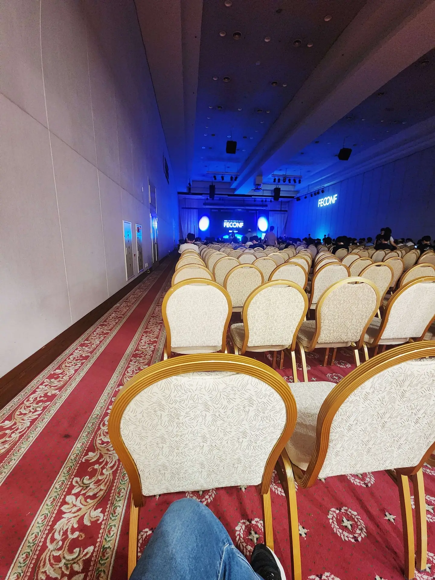 FEConf 행사장 내부 전경. 붉은 카펫이 깔린 넓은 홀의 맨 뒤쪽 자리에서 무대를 향해 찍은 사진으로, 무대 중앙 스크린에 ‘FEConf’ 로고가 파란 조명 속에 빛나고 있다. 객석에는 연한 베이지색 의자들이 줄지어 놓여 있으며, 일부 관객이 자리에 앉아 있는 모습이 보인다.